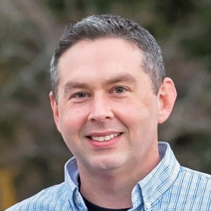 Bob wearing a blue dress shirt standing in front of a tree